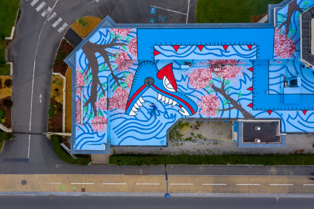 Close vertical aerial view of Oré's Quetzalcoatl mural on the roof of the Pavillon des Fleurs, Bagnoles-de-l'Orne, showing the serpent's open-jawed head in blue, red, and white against a wave-patterned background, 2022.
