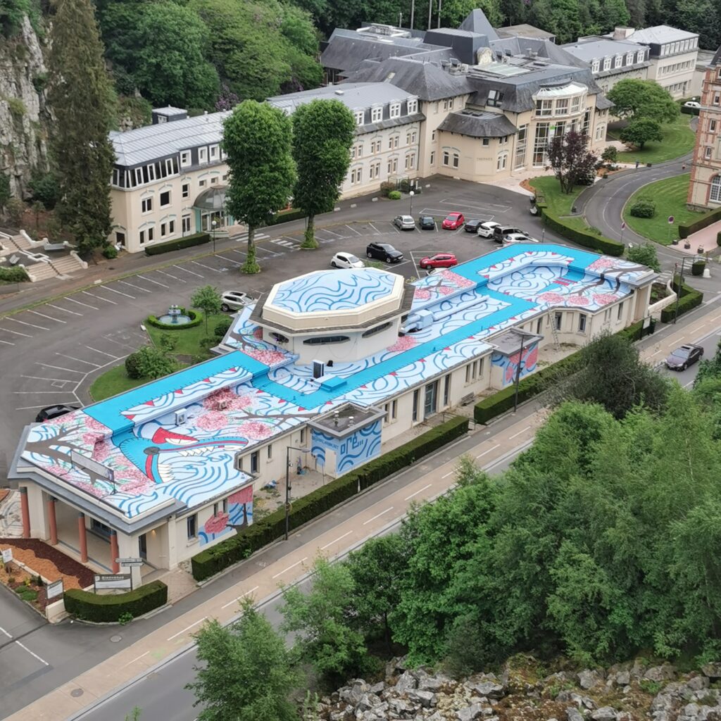 Aerial oblique view of the Pavillon des Fleurs thermal spa, Bagnoles-de-l'Orne, showing Oré's Quetzalcoatl mural covering the entire roof of the building, seen from the Roc au Chien viewpoint, 2022.