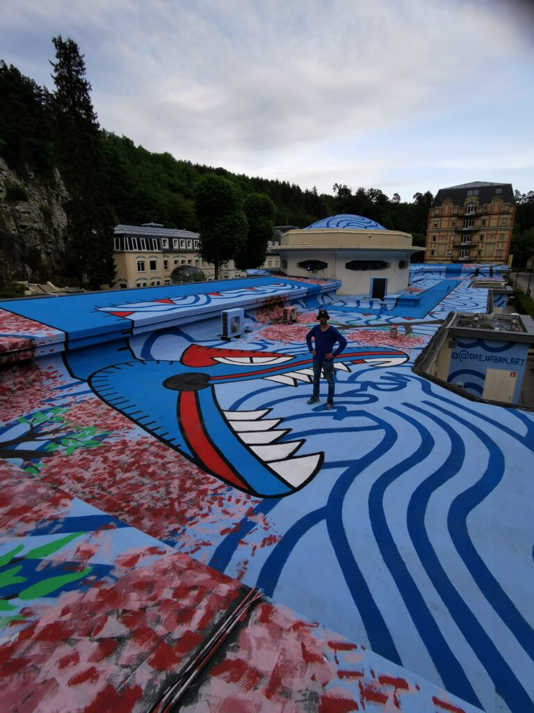Artist Oré standing on the roof of the Pavillon des Fleurs beside the head of his Quetzalcoatl mural, Bagnoles-de-l'Orne, 2022, illustrating the monumental scale of the work.